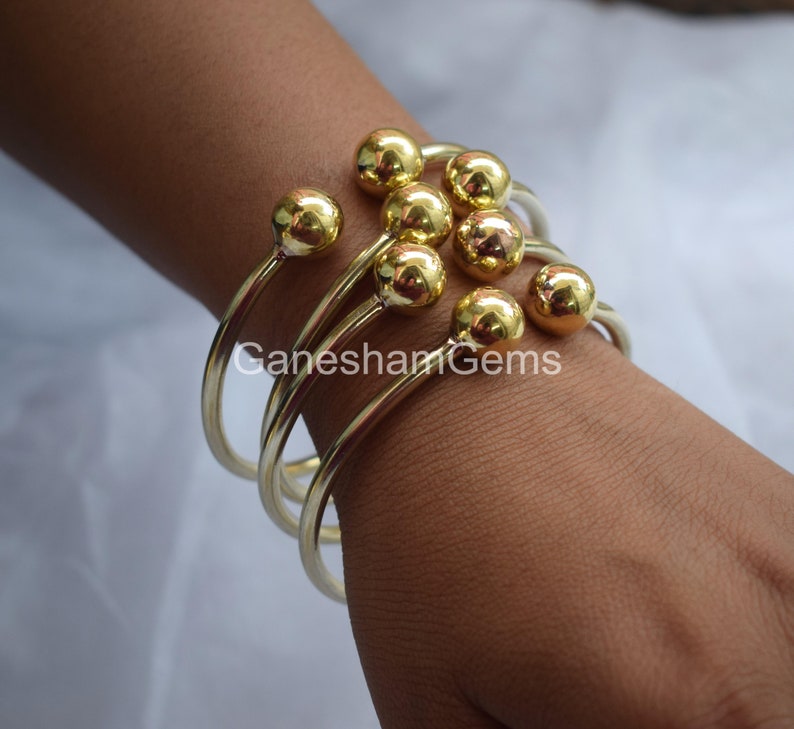 May include: Gold bangle bracelets with round ball accents. The bracelets are stacked on a person's wrist.
