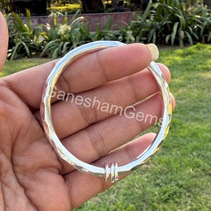 Pode incluir: Uma pulseira de prata martelada com três pequenos anéis. A pulseira tem uma superfície texturizada e é segurada em uma mão. O texto "Ganesham Gems" é visível na pulseira. O fundo está desfocado com grama verde e folhagem.