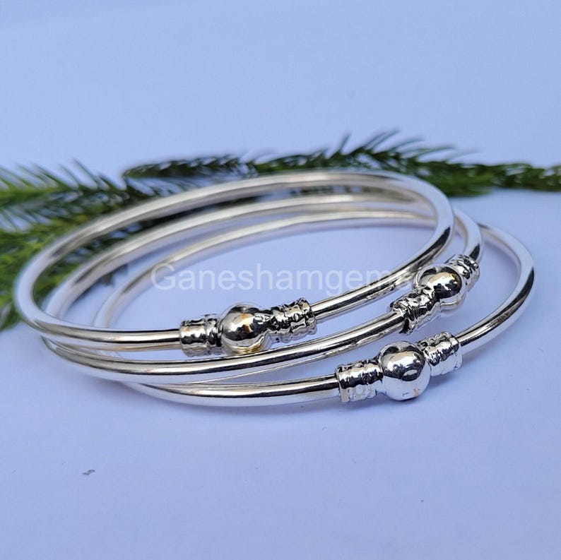 Peut inclure: Ensemble de trois bracelets argent&eacute;s. Chaque bracelet pr&eacute;sente une perle sph&eacute;rique centrale et des d&eacute;tails d&eacute;coratifs. Les bracelets sont empil&eacute;s, avec une brindille de verdure en arri&egrave;re-plan. Le texte "Ganeshamgems" est visible.