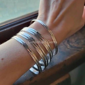 May include: A collection of silver bangles with a hammered texture are stacked on a wrist. The bangles are made of a shiny, metallic material and reflect light. The jewelry is displayed on a person's arm.