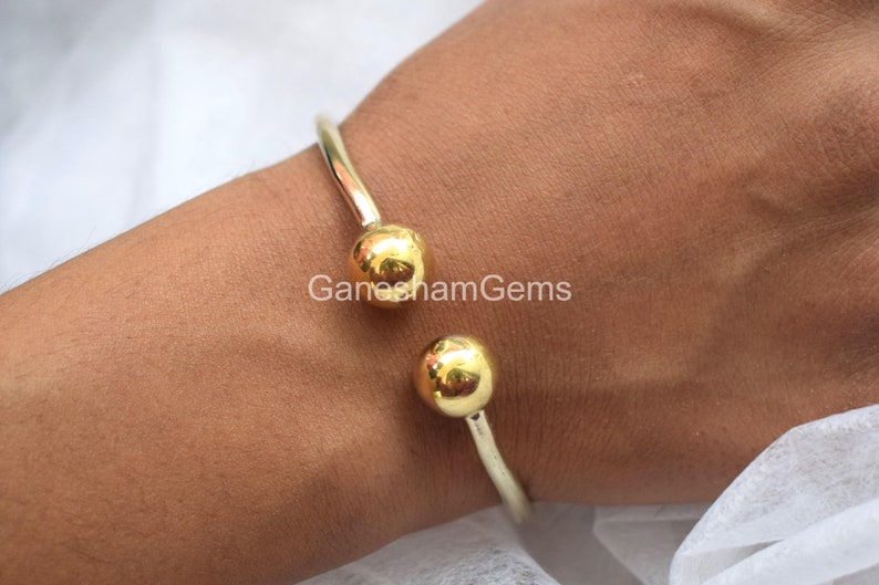 May include: A gold bangle bracelet with two round, gold balls at the ends. The bracelet is open and can be adjusted to fit different wrist sizes.