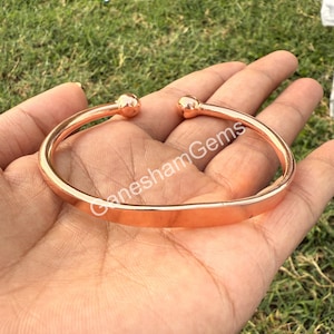 May include: A rose gold cuff bracelet with rounded ball ends. The bracelet is open and sits on a hand. The text "GaneshamGems" is visible on the bracelet.