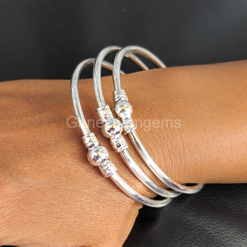 Peut inclure: Trois bracelets argent&eacute;s avec des accents de perles d&eacute;coratives sont pr&eacute;sent&eacute;s sur un bras. Les bracelets sont incurv&eacute;s et ont une finition lisse et polie. Les perles sont rondes et carr&eacute;es, ajoutant un int&eacute;r&ecirc;t visuel aux bijoux.