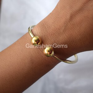 May include: A gold bangle bracelet with two round balls on the ends. The bracelet is on a person's wrist.