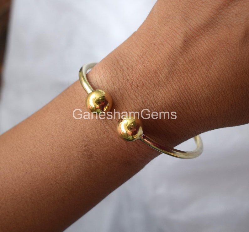 May include: A gold bangle bracelet with two round balls on the ends. The bracelet is on a person's wrist.