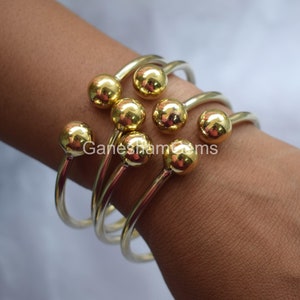 May include: Set of five gold-toned bangle bracelets with round ball accents. The bracelets are stacked on a person's wrist.
