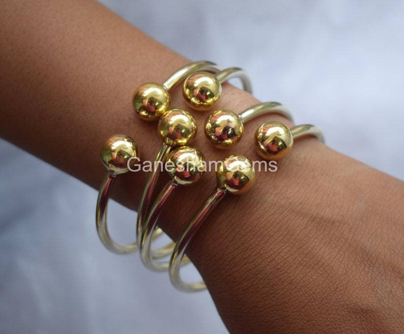 May include: Set of five gold-toned bangle bracelets with round ball accents. The bracelets are stacked on a person's wrist.