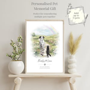 Op de afbeelding: Een ingelijste print met een Dalmatische hond en een zwarte kat die op een pad lopen, met een regenboog op de achtergrond. De tekst luidt "Personalised Pet Memorial Gift" en "Freckles & Lyna". De print staat op een houten plank.