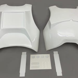 Könnte beinhalten: Weißes Brust- und Rückenpanzer aus Kunststoff sowie zusätzliche Komponenten. Die Rüstung hat eine glänzende Oberfläche und ein rechteckiges Detail auf dem Brustteil. Enthält zwei Riemen und einen Bogen mit Aufklebern.