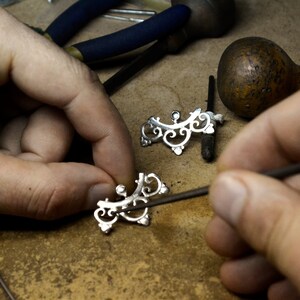 May include: A close-up of a jeweler working on a silver filigree design. The jeweler is using a small tool to shape the delicate metalwork.