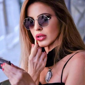 May include: A woman wearing a black top with a silver necklace featuring a large, oval-shaped stone pendant. She is applying pink lipstick while looking in a small mirror.