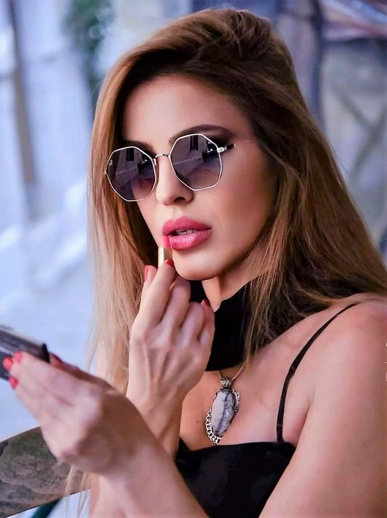 May include: A woman wearing a black top with a silver necklace featuring a large, oval-shaped stone pendant. She is applying pink lipstick while looking in a small mirror.