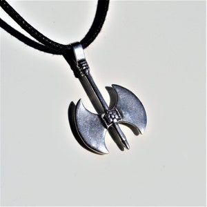 Può includere: Un ciondolo a forma di ascia a doppia testa in argento su un cordino nero. L'ascia presenta un design dettagliato con un piccolo motivo circolare nella parte superiore del manico.