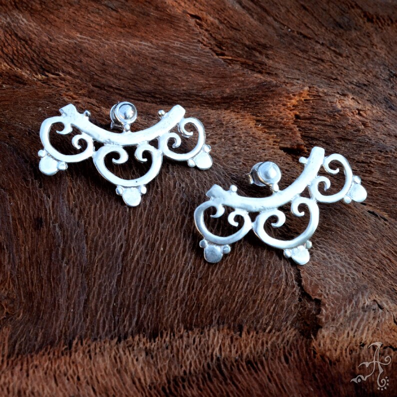 May include: A pair of silver filigree earrings with a delicate, intricate design. The earrings are shaped like a crescent moon and have a small, round bead at the top.