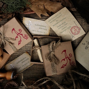 May include: A close-up of a brown box with a red and white logo that says "Alexandros Kontaxis Art Jewelry". The box is tied with twine and has a small, red, wax seal. The box is sitting on a burlap surface.