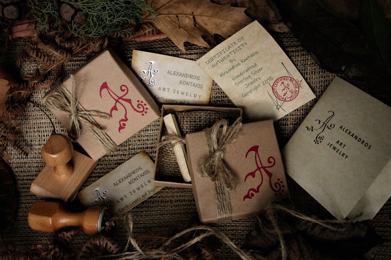 May include: A close-up of a brown box with a red and white logo that says "Alexandros Kontaxis Art Jewelry". The box is tied with twine and has a small, red, wax seal. The box is sitting on a burlap surface.