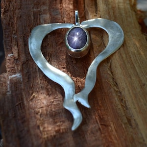 Può includere: Ciondolo in argento a forma di cuore, con una pietra ovale viola scuro. Il ciondolo è presentato su uno sfondo di legno, evidenziando il design e la maestria del gioiello.