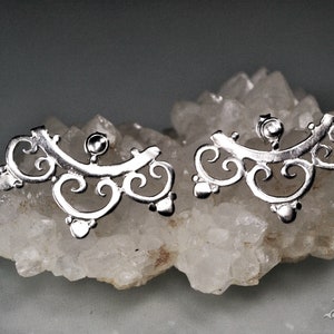 May include: A pair of silver earrings with a delicate, intricate design. The earrings are shaped like a crescent moon with a series of swirls and curves.