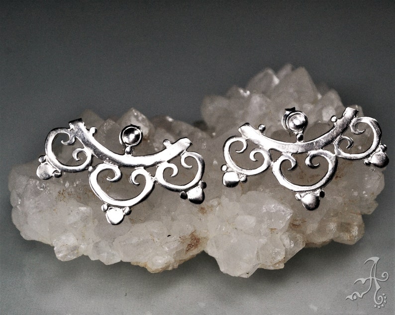 May include: A pair of silver earrings with a delicate, intricate design. The earrings are shaped like a crescent moon with a series of swirls and curves.
