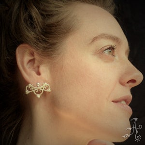 May include: A pair of silver ear cuffs with a delicate, ornate design. The cuffs are shaped like a crescent moon and feature intricate swirls and curves. The cuffs are worn by a person with long brown hair.