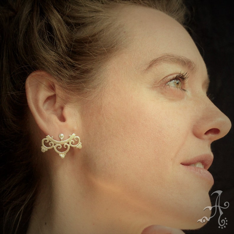 May include: A pair of silver ear cuffs with a delicate, ornate design. The cuffs are shaped like a crescent moon and feature intricate swirls and curves. The cuffs are worn by a person with long brown hair.