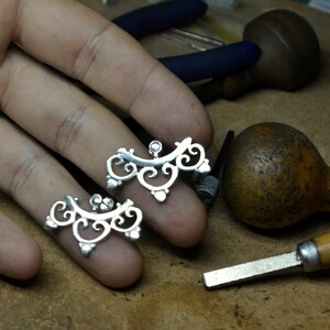 May include: Two silver filigree earrings with a delicate, intricate design. The earrings are shown on a hand, with a wooden surface and tools in the background.