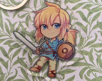 May include: A clear acrylic keychain featuring a cartoon illustration of Link from the Legend of Zelda video game series. Link is depicted in a blue tunic, holding a sword and shield, with blonde hair and blue eyes.