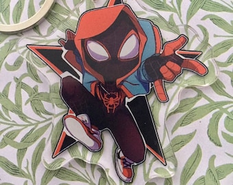 May include: A red and black cartoon Spider-Man character with a blue jacket, wearing white sneakers, is featured on a star-shaped keychain. The keychain is made of clear acrylic.
