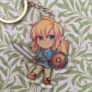 Puede incluir: Un llavero de acrílico transparente con una ilustración de dibujos animados de Link de la serie de videojuegos The Legend of Zelda. Link está representado con una túnica azul, sosteniendo una espada y un escudo, con cabello rubio y ojos azules.
