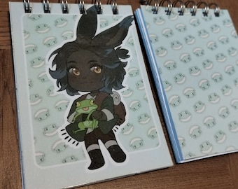 May include: A blue and white patterned notebook with a sticker of a cartoon character with black hair, brown skin, and a green frog. The character is wearing a brown shirt, black pants, and white boots.