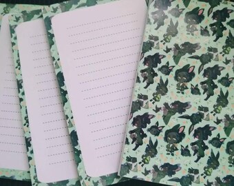 May include: A set of four notebooks with lined pages. The covers feature a repeating pattern of cartoon characters on a light green background. The edges of the pages have a green camouflage design.