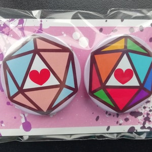 Può includere: Due bottoni da spilla da 2,5 cm di diametro con un design geometrico arcobaleno e pastello. Ogni bottone presenta un triangolo bianco con un cuore rosso al centro. I bottoni sono su uno sfondo rosa e bianco con schizzi di vernice viola.