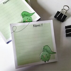 Op de afbeelding: Een stapel groen en wit gestreept notitieboekpapier met een cartoonfiguur op elke pagina. Het figuur is een groen wezen met een blad op zijn hoofd. Het notitieboek heeft de tekst "Notes!!" op de voorkant.