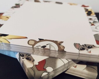 May include: A stack of white notepaper with a border of cartoon animals in various poses. The animals include a bear, a husky, and a bird, all with festive attire. The paper is stacked on a dark surface.