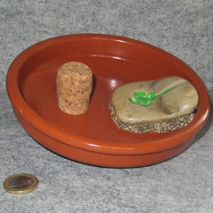 May include: A terracotta-colored, oval-shaped dish with a cork stopper and a decorated stone. The stone has a green clover design. A euro coin is visible in the foreground. The dish is likely for decorative purposes.