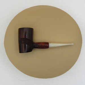 May include: A dark brown tobacco pipe with a light-coloured stem. The pipe has a textured, dark brown bowl and a reddish-brown section connecting to the stem. The stem is a light cream colour. The pipe is set against a tan circular background.