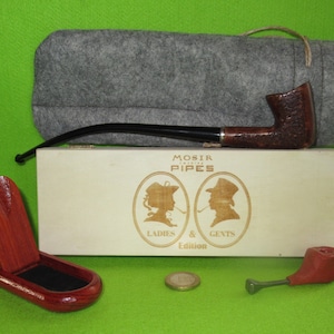 Pode incluir: Uma caixa de madeira com um cachimbo marrom e uma bolsa de feltro. A caixa tem as palavras "Mosir Pipes Ladies & Gents Edition" impressas nela. O cachimbo está em uma bolsa de feltro marrom.