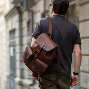Könnte beinhalten: Ein brauner Lederrucksack mit mehreren Taschen und einem Klappenverschluss wird über der Schulter getragen. Die Person trägt ein dunkelgraues T-Shirt, olivgrüne Shorts und Turnschuhe. Das Bild wird im Freien auf einer Kopfsteinpflasterstraße aufgenommen.
