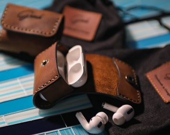 May include: Two brown leather cases for wireless earbuds, one open with white earbuds inside. The cases have the word "Good" embossed on them. The cases are on a blue and white tiled surface.