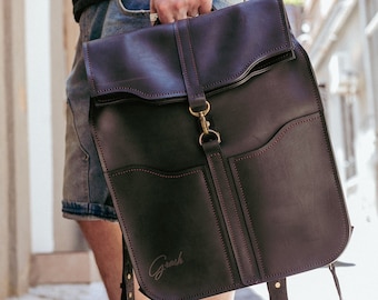 Könnte beinhalten: Ein dunkelbrauner Lederrucksack mit roten Nähten und einem Messingverschluss. Der Rucksack hat zwei große Taschen auf der Vorderseite und eine kleinere Tasche oben. Das Wort "Grach" ist auf der Vorderseite des Rucksacks geprägt.