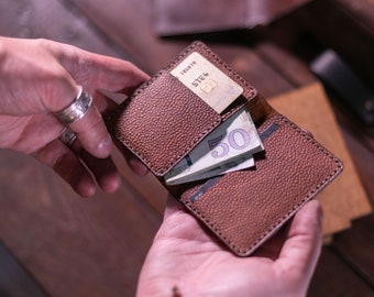May include: Brown leather wallet with a textured finish, open to reveal a credit card and a stack of paper currency. The wallet is held open, showcasing its interior design and storage capacity. The stitching is visible.