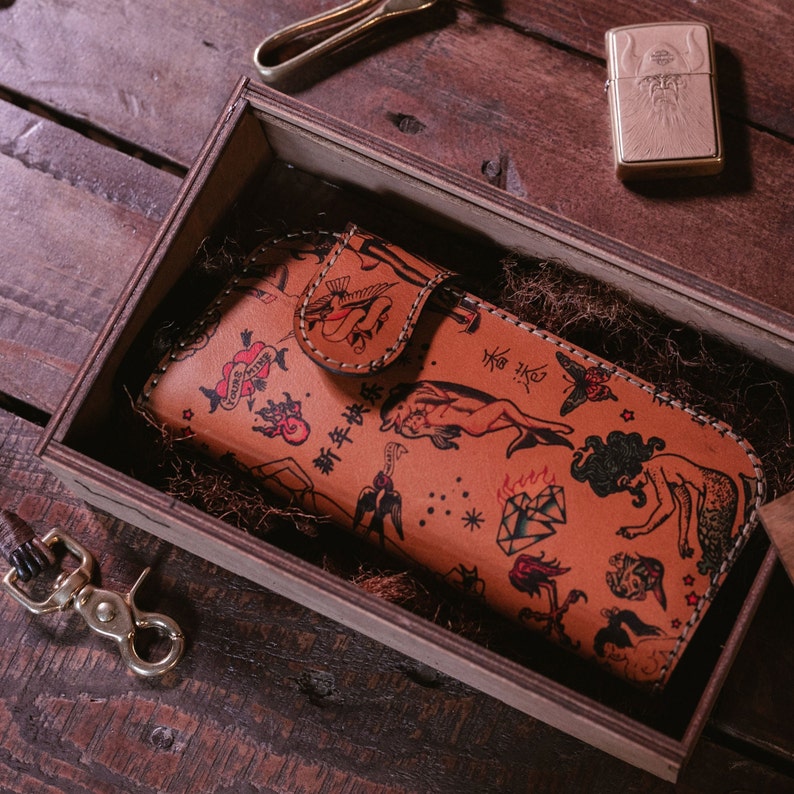 May include: A brown leather wallet with a flap closure and a colorful tattoo-style design. The wallet is filled with a variety of colorful tattoos, including hearts, stars, birds, and flowers. The wallet is sitting in a wooden box.