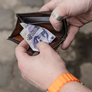 Op de afbeelding: Een bruine leren portemonnee met een ritssluiting is open en toont een Tsjechisch bankbiljet van 100 kronen.