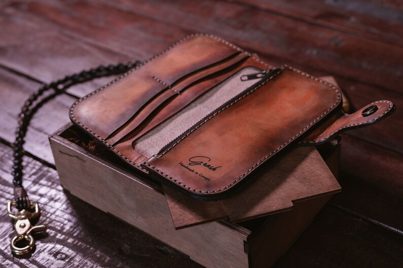 May include: A brown leather wallet with a chain attached. The wallet is open and has a zippered pocket and multiple card slots. The wallet is embossed with the text "Greesh Handmade in Georgia".