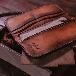 May include: A brown leather wallet with a zippered pocket and a chain attached. The wallet is open and the inside is lined with a light brown fabric. The wallet has the text "Crush Handmade in Germany" embossed on the front.