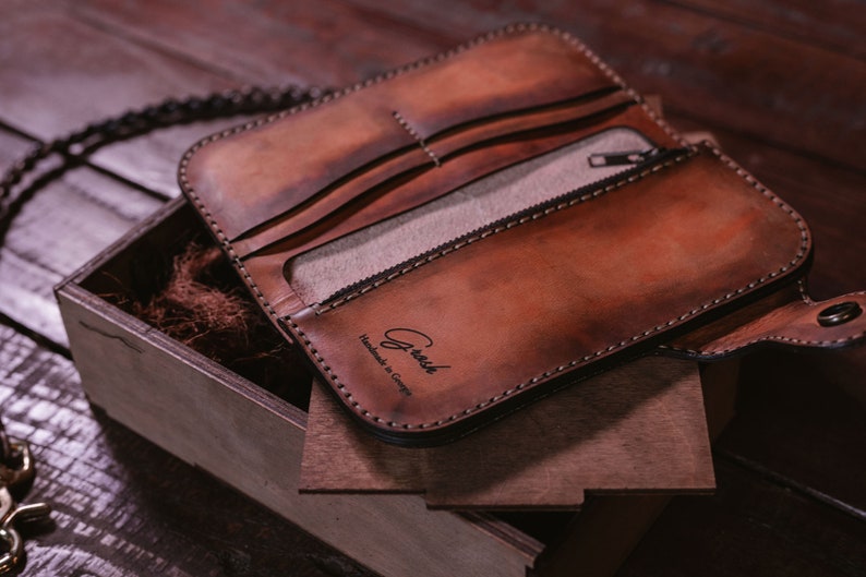 May include: A brown leather wallet with a zippered pocket and a chain attached. The wallet is open and the inside is lined with a light brown fabric. The wallet has the text "Crush Handmade in Germany" embossed on the front.