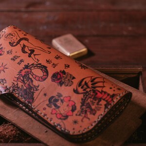May include: A brown leather wallet with a detailed design of various tattoos, including a dragon, a snake, a woman, and a heart. The wallet is folded and has a stitched edge.