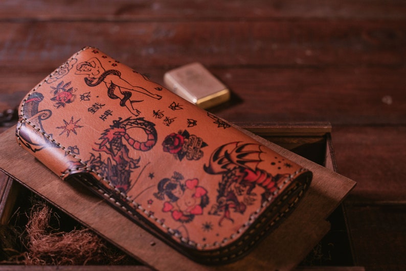 May include: A brown leather wallet with a detailed design of various tattoos, including a dragon, a snake, a woman, and a heart. The wallet is folded and has a stitched edge.