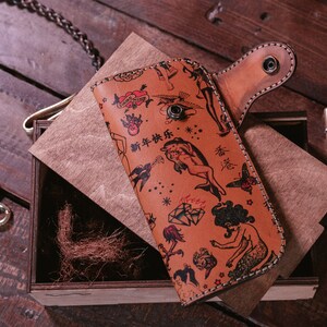 May include: A brown leather wallet with a flap closure and a snap button. The wallet is decorated with a colorful tattoo-style pattern featuring images of mermaids, hearts, stars, and the text "新年快乐".
