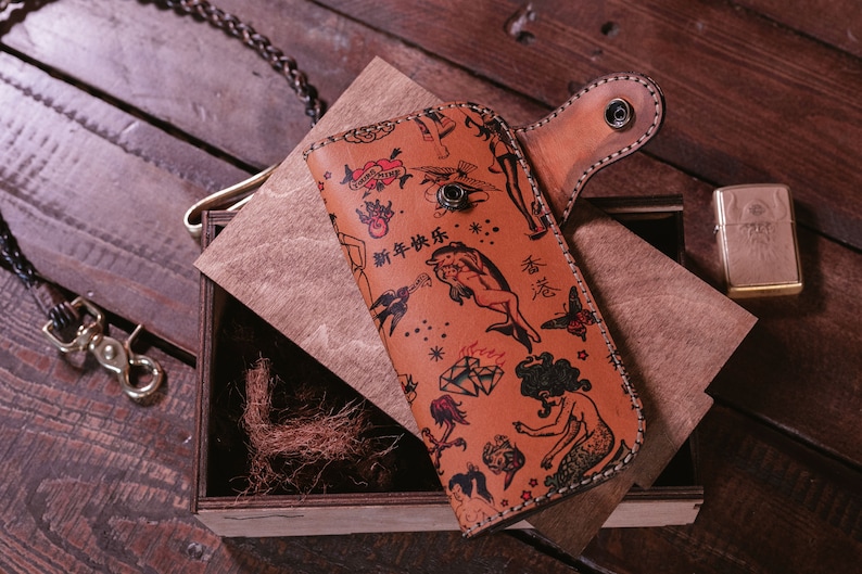 May include: A brown leather wallet with a flap closure and a snap button. The wallet is decorated with a colorful tattoo-style pattern featuring images of mermaids, hearts, stars, and the text "新年快乐".